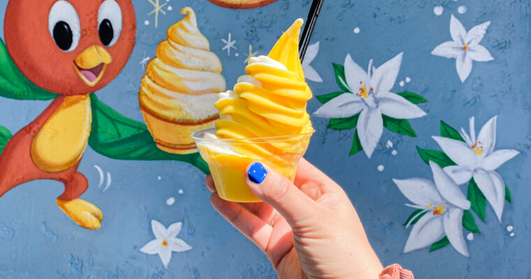 Hand holding a soft-serve Dole Whip frozen treat with Orange Bird art in the background in Magic Kingdom at Walt Disney World.